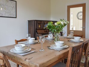 Uma mesa de jantar posta com tigelas, pratos e copos de vinho brancos, um vaso com flores e um piano de madeira ao fundo.