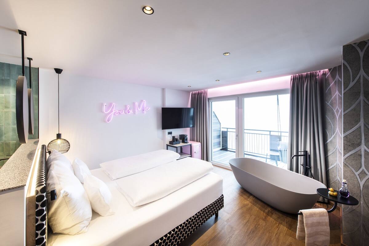 Une chambre d’hôtel moderne avec un grand lit, une baignoire, un balcon avec des rideaux et un panneau lumineux au néon.
