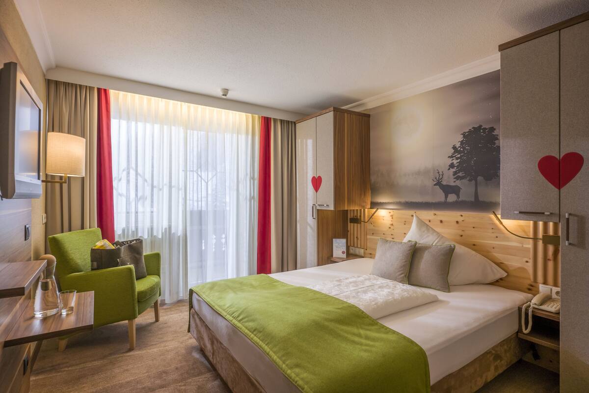 Une chambre d’hôtel avec un lit, un fauteuil vert, un bureau avec une lampe et un tableau représentant un cerf accroché au mur.