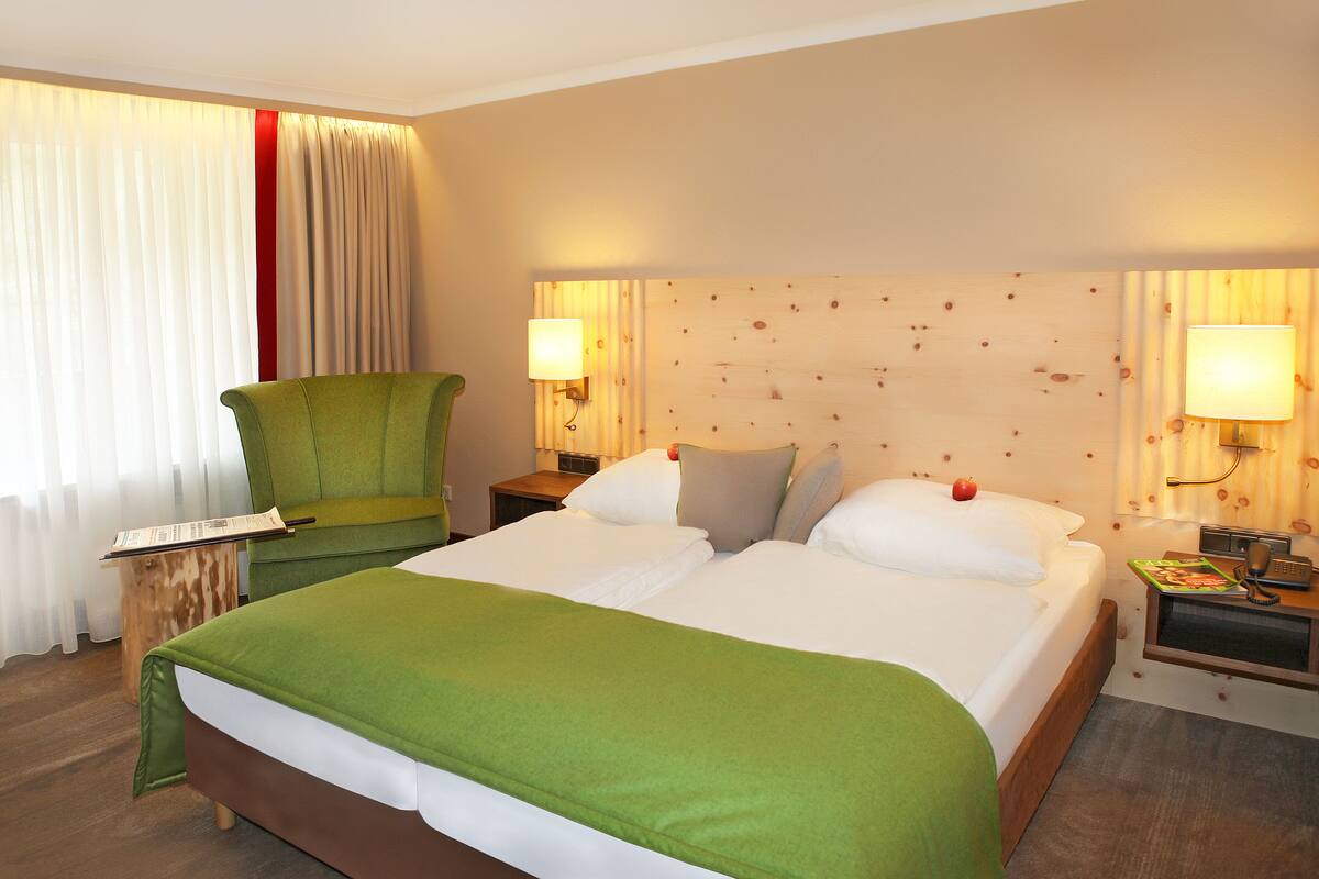 Une chambre d’hôtel avec un grand lit, un fauteuil vert, deux tables de chevet avec des lampes et une fenêtre avec des rideaux.