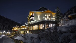 Een groot, goed verlicht hotel met meerdere balkons, een rivier op de voorgrond en met sneeuw bedekte bomen.