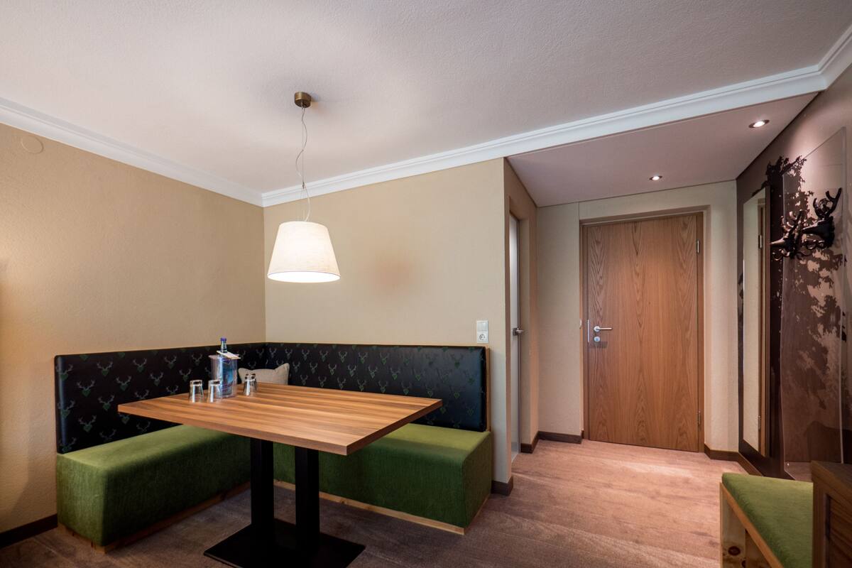Une pièce avec une table en bois, des sièges rembourrés de couleur verte, une lampe suspendue et une porte en bois.