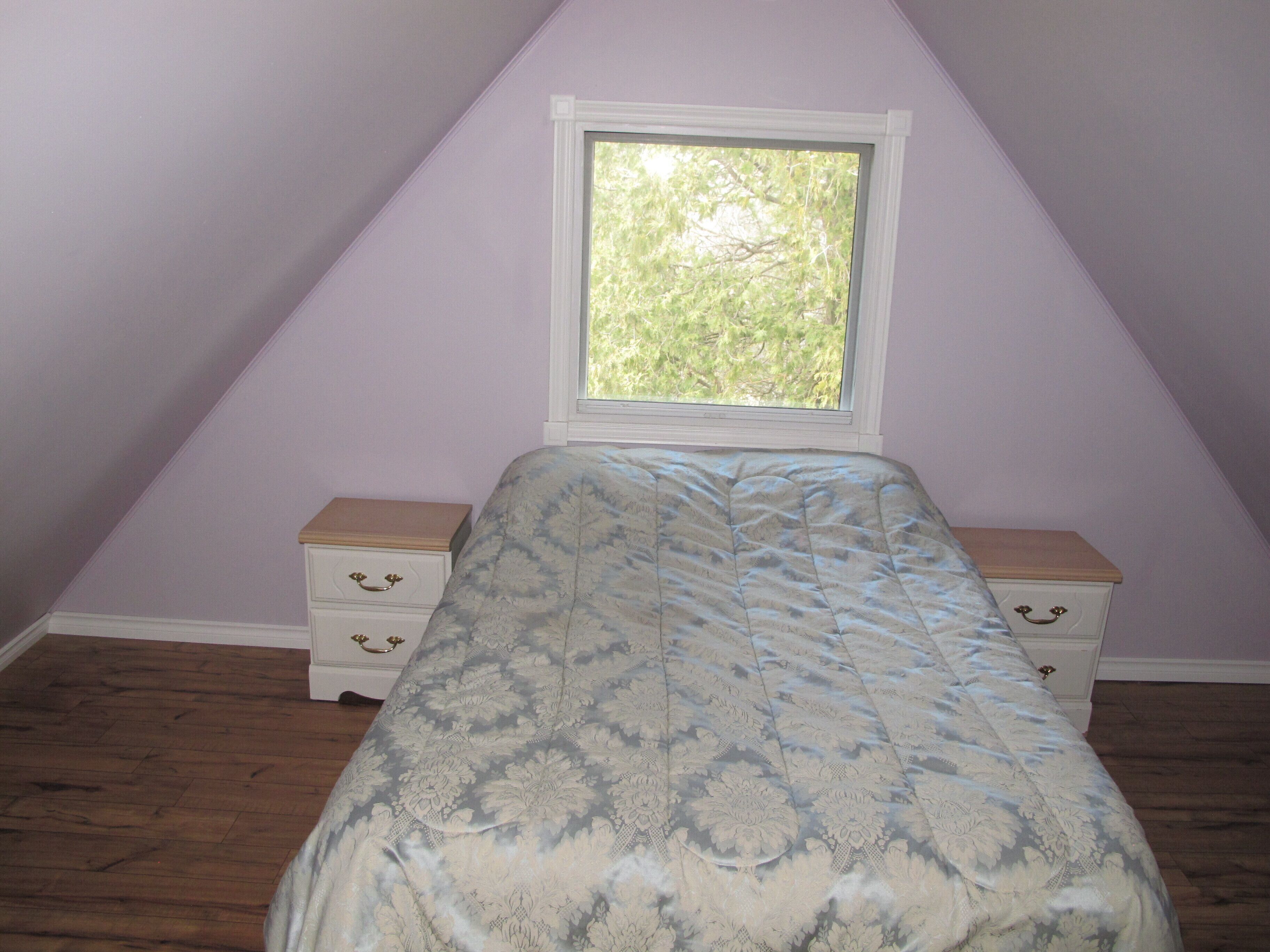 A bedroom with a sloped ceiling, a window with a view of trees, a bed with a patterned bedspread, and two bedside tables with drawers.