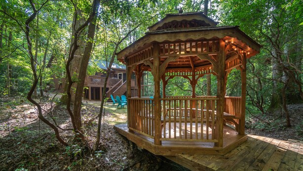 A wooden gazebo with a wooden deck, surrounded by a forested area.