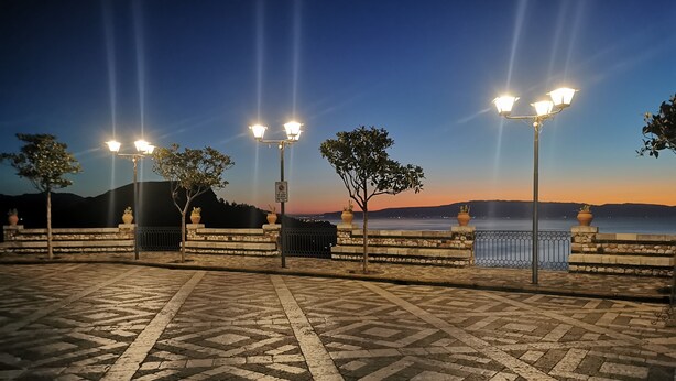 Ein gepflasterter Weg mit Steinplatten und Straßenlaternen, der zu einem Gewässer mit Sonnenuntergang im Hintergrund führt.