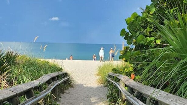 Ein hölzerner Steg führt zu einem Strand mit blauem Himmel und ruhiger See.