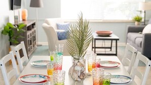 Ein Esstisch ist gedeckt mit buntem Glas und Geschirr, ein dekoratives Mittelstück und ein moderner Wohnbereich im Hintergrund.