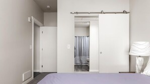 Ein Schlafzimmer mit Bett, Nachttisch, Lampe, Kleiderschrank und einem durchgegangenen Tür sichtbar werdenden Badezimmer.
