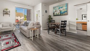 Ein modernes Wohnzimmer mit Sofa, Couchtisch und Essbereich. Ein Gemälde hängt an der Wand und es gibt einen Blick ins Freie.