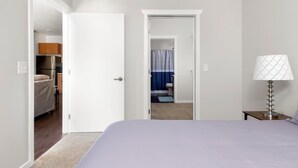 A hotel room with a bed, a nightstand with a lamp, a door leading to a bathroom, and a view of a kitchen area. - *NEW* Spacious Condo Near Downtown TC (Traverse City)