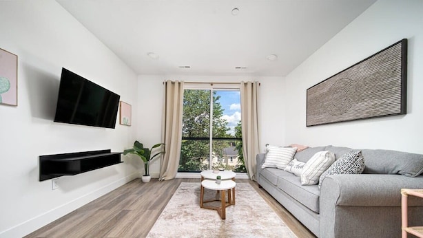 A modern living room with a grey sofa, a flat-screen TV, a coffee table, and a large window with curtains. - 1BR I King + Sofa I Walk Shops + DT TC (Traverse City)