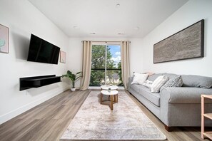 Ein modernes Wohnzimmer mit grauem Sofa, einem Flachbildfernseher, einem Couchtisch und einem großen Fenster mit Vorhängen.