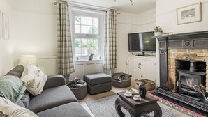 A living room with a sofa, ottoman, fireplace, television, and a window with curtains. - 4 bedroom accommodation in Bacton, Norwich (Bacton, Norwich)