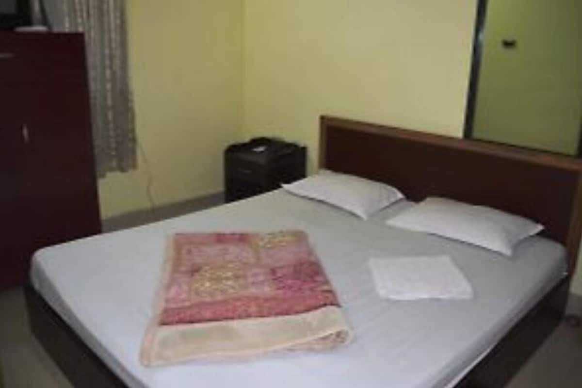 Un lit simple avec un drap blanc et une couverture rose, une tête de lit en bois et une petite table de chevet sur laquelle se trouve un objet noir.