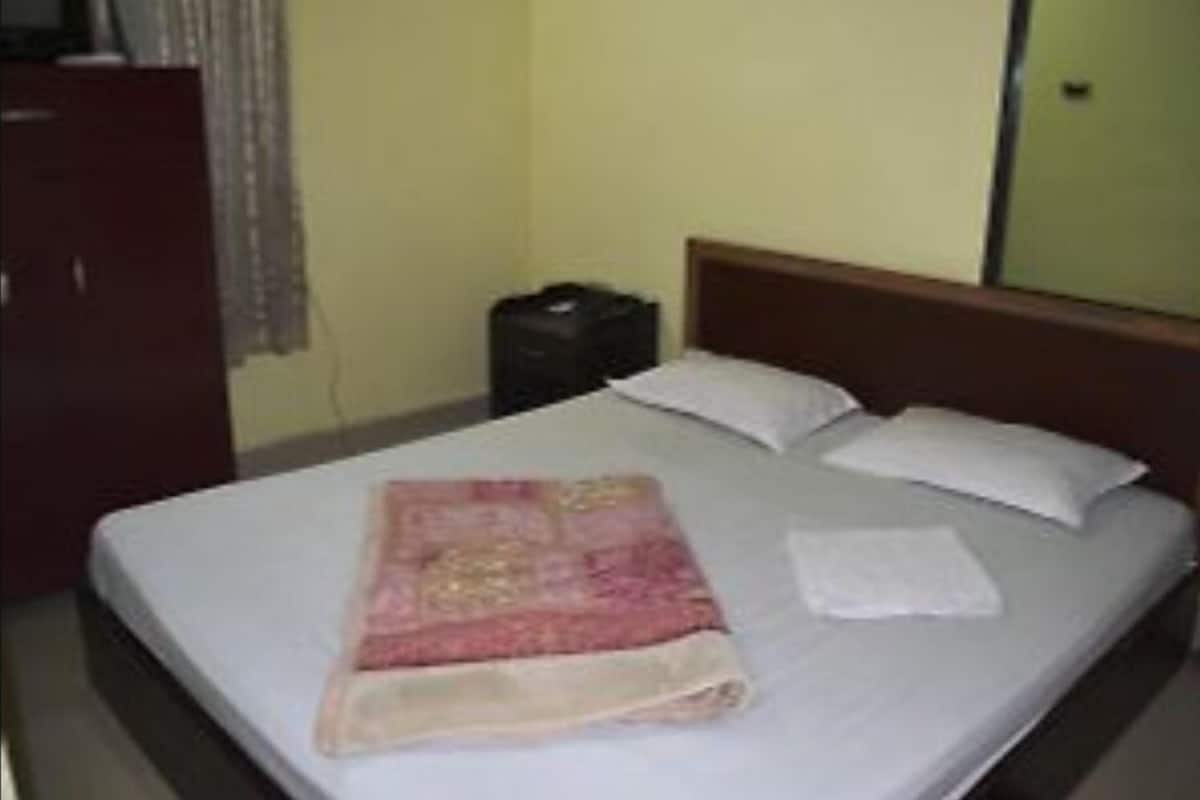 Un lit simple avec un matelas blanc et deux oreillers, une table de chevet avec un sac noir, et une tête de lit en bois.