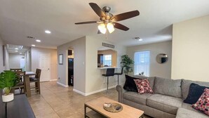 A spacious living room with a sofa, coffee table, and a ceiling fan. There is a dining area with chairs and a table, and a kitchen in the background. - Quarry Downtown Airport 10 mins or less (San Antonio)