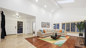 Un salon spacieux avec un grand canapé d’angle, une table basse et un tapis coloré. Il y a deux portes qui donnent sur d’autres pièces, ainsi qu’une grande fenêtre avec des stores.