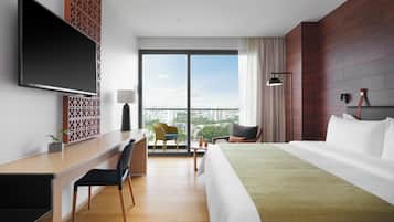 A modern hotel room with a large bed, a desk with a chair, a flat-screen TV, and a view of the city through large windows.