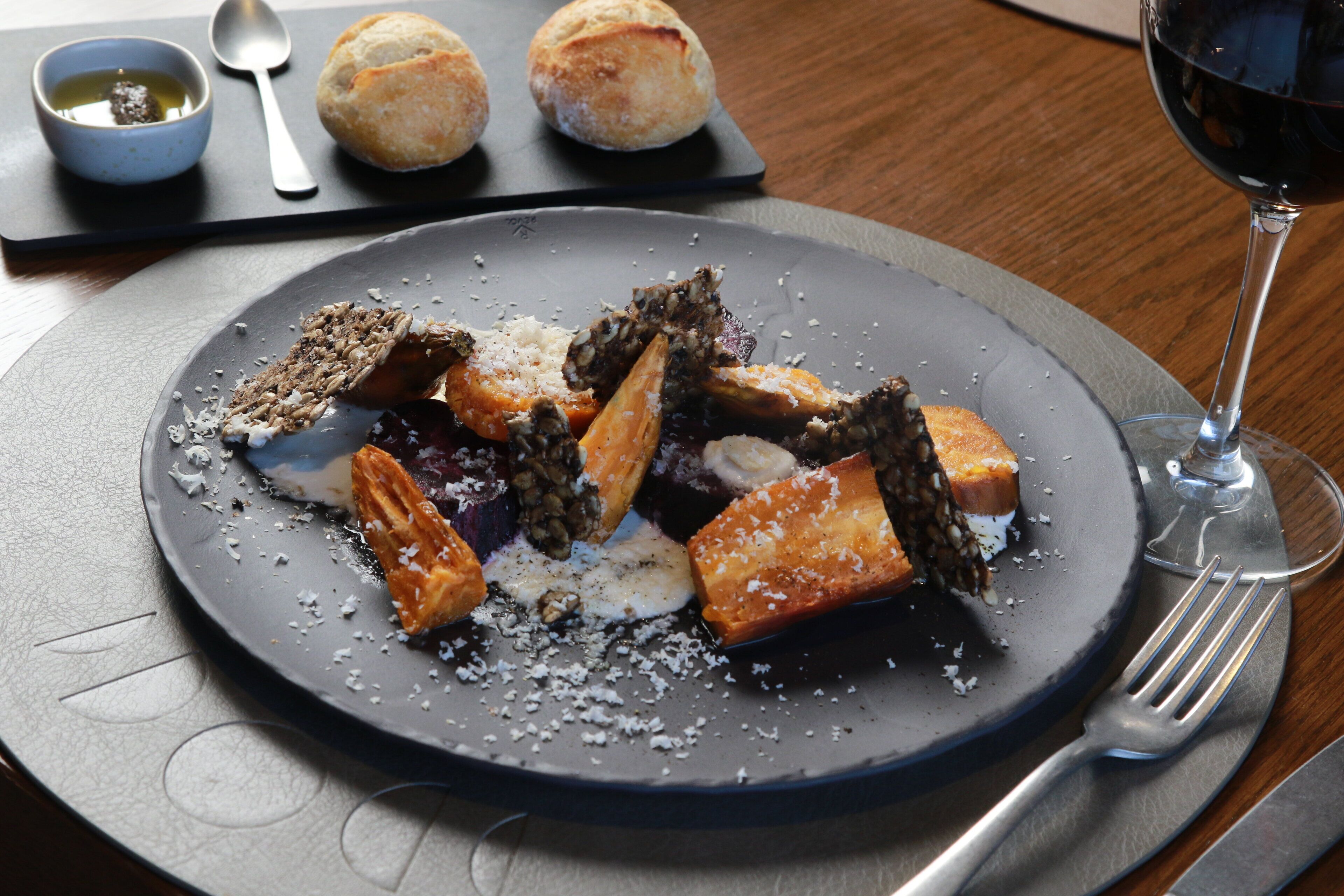 A plate of food with various items including bread rolls, a bowl of sauce, and a glass of red wine.