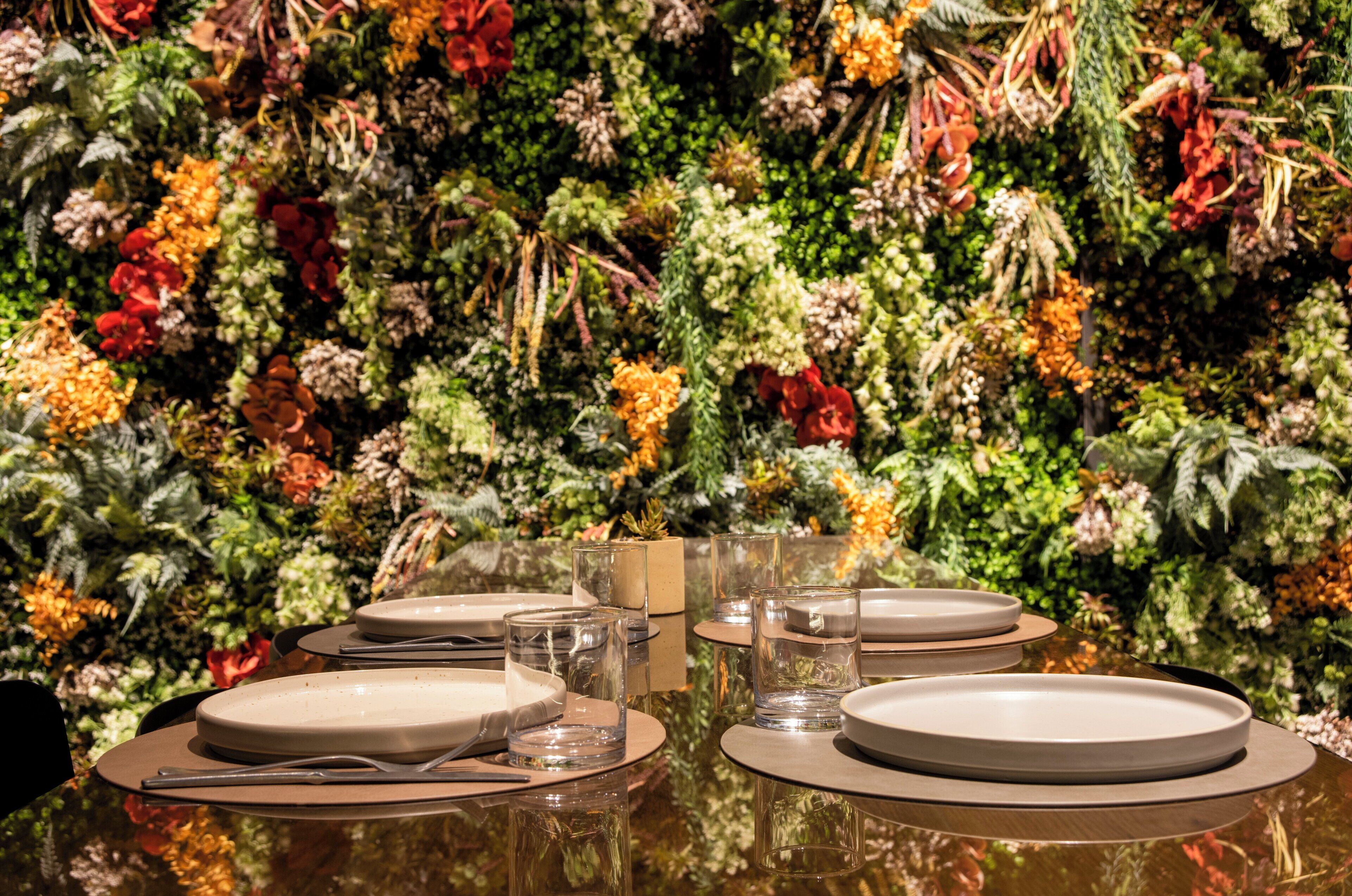 A dining table set with plates, glasses, and cutlery against a backdrop of a lush, green wall adorned with various plants and flowers.
