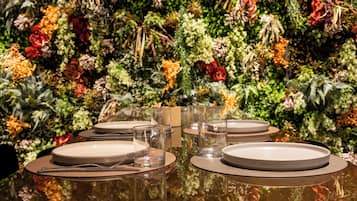 A dining table set with plates, glasses, and cutlery against a backdrop of a lush, green wall adorned with various plants and flowers.