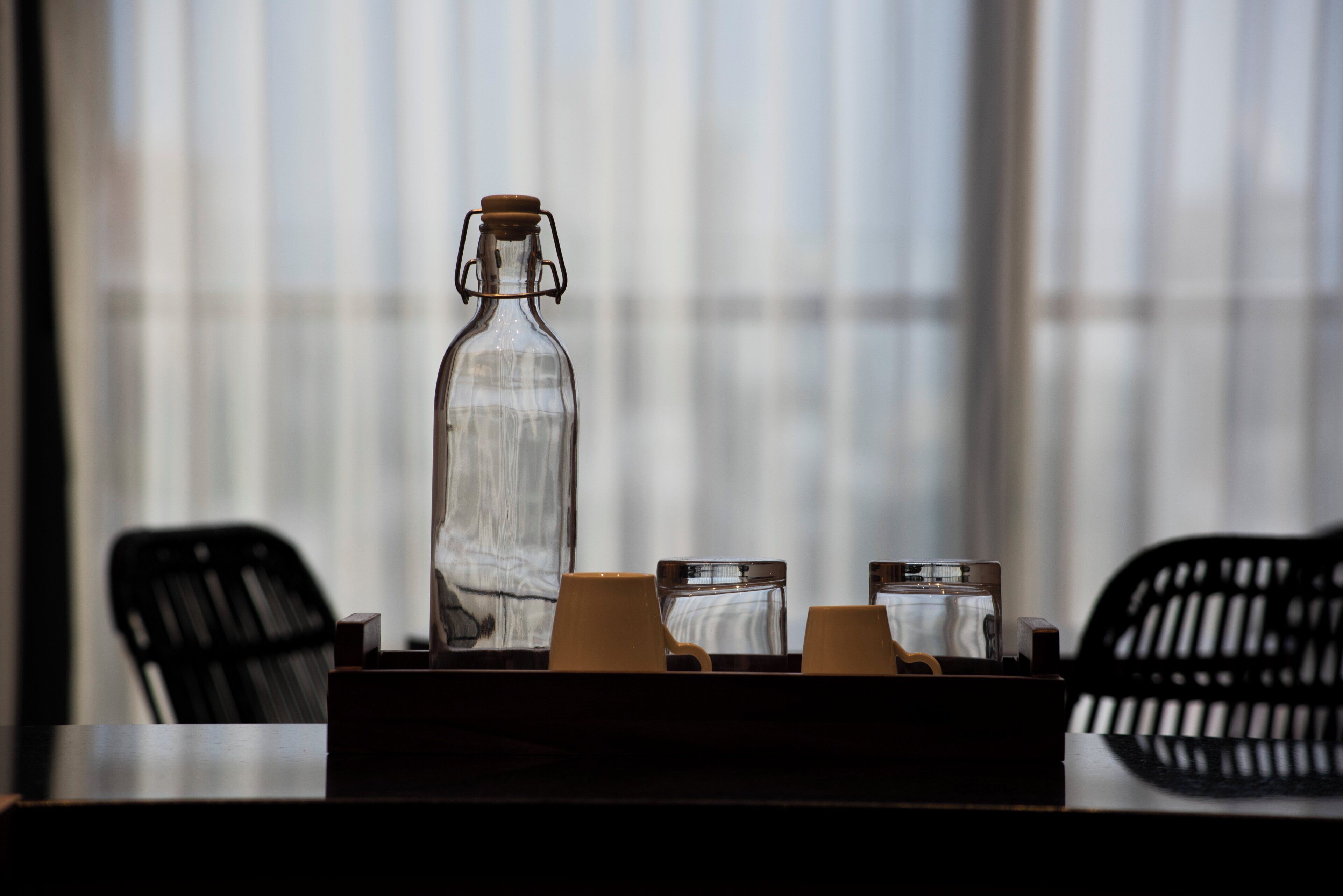 A clear glass bottle on a table with two cups and a small box, set against a window with blinds.