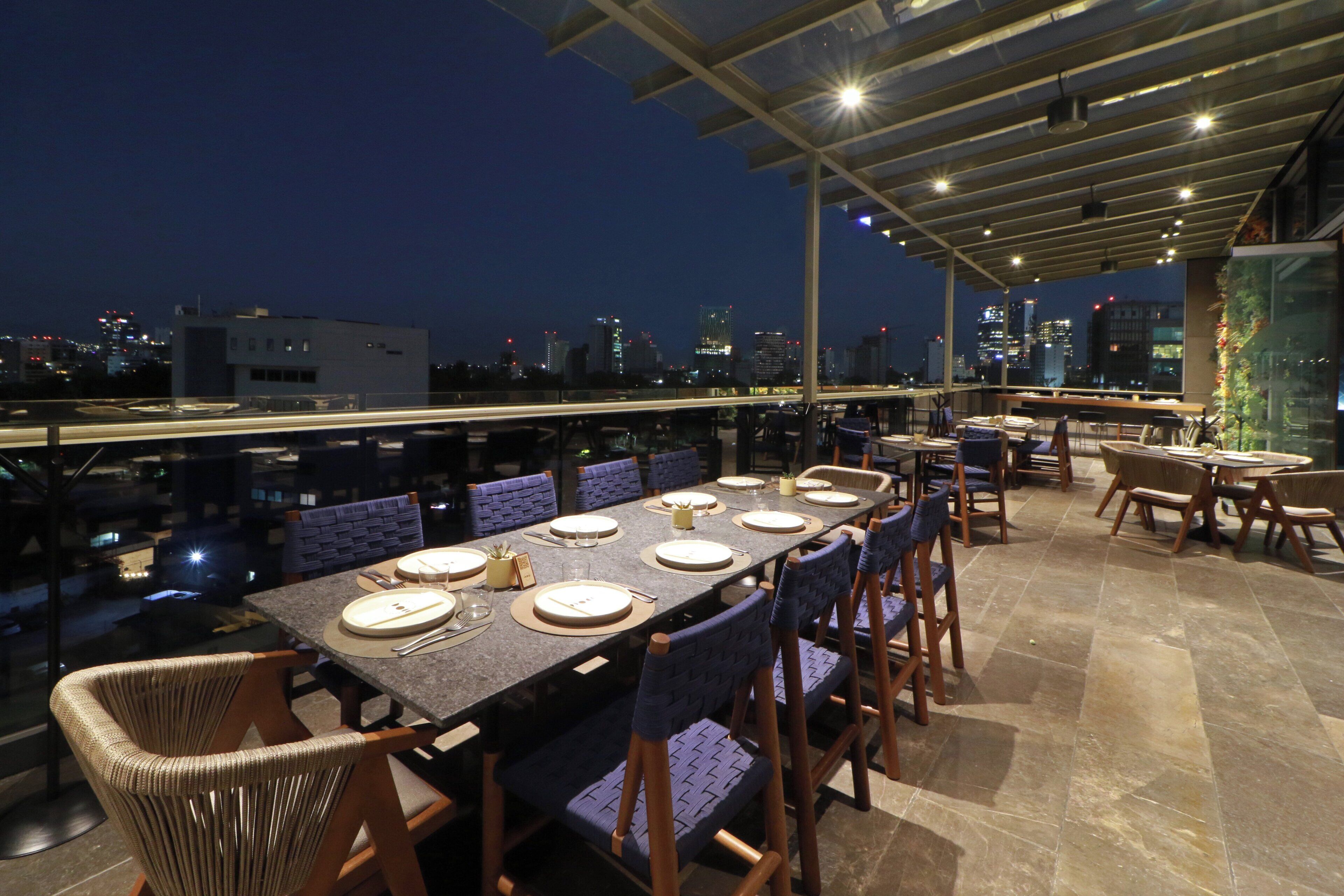 Un área de comedor en la azotea con una mesa larga dispuesta para seis personas, rodeada de sillas, y una vista panorámica de la ciudad al fondo.