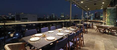 Un área de comedor en la azotea con una mesa larga dispuesta para seis personas, rodeada de sillas, y una vista panorámica de la ciudad al fondo.