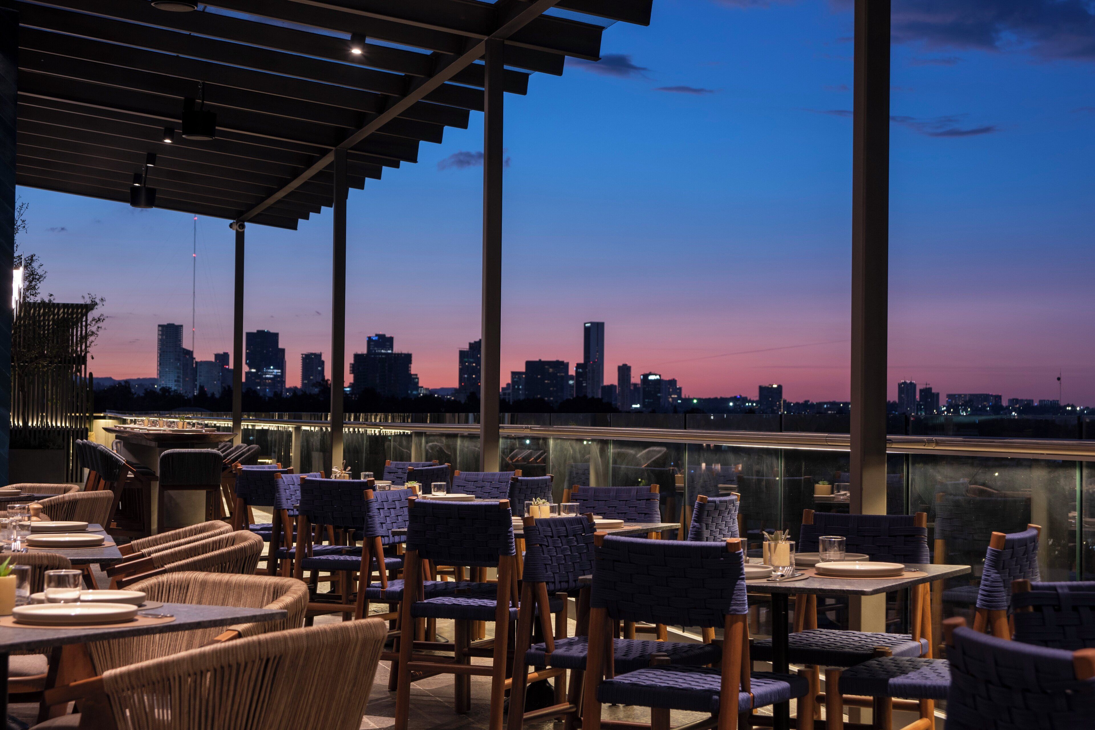 Un área de comedor al aire libre con sillas y mesas de mimbre, con vistas a un horizonte urbano al atardecer.