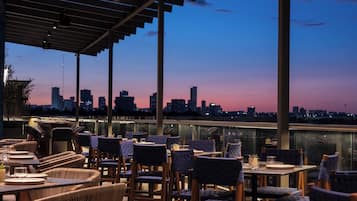 Un área de comedor al aire libre con sillas y mesas de mimbre, con vistas a un horizonte urbano al atardecer.
