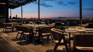 Un área de comedor al aire libre con mesas y sillas, con vistas a un horizonte urbano al atardecer.