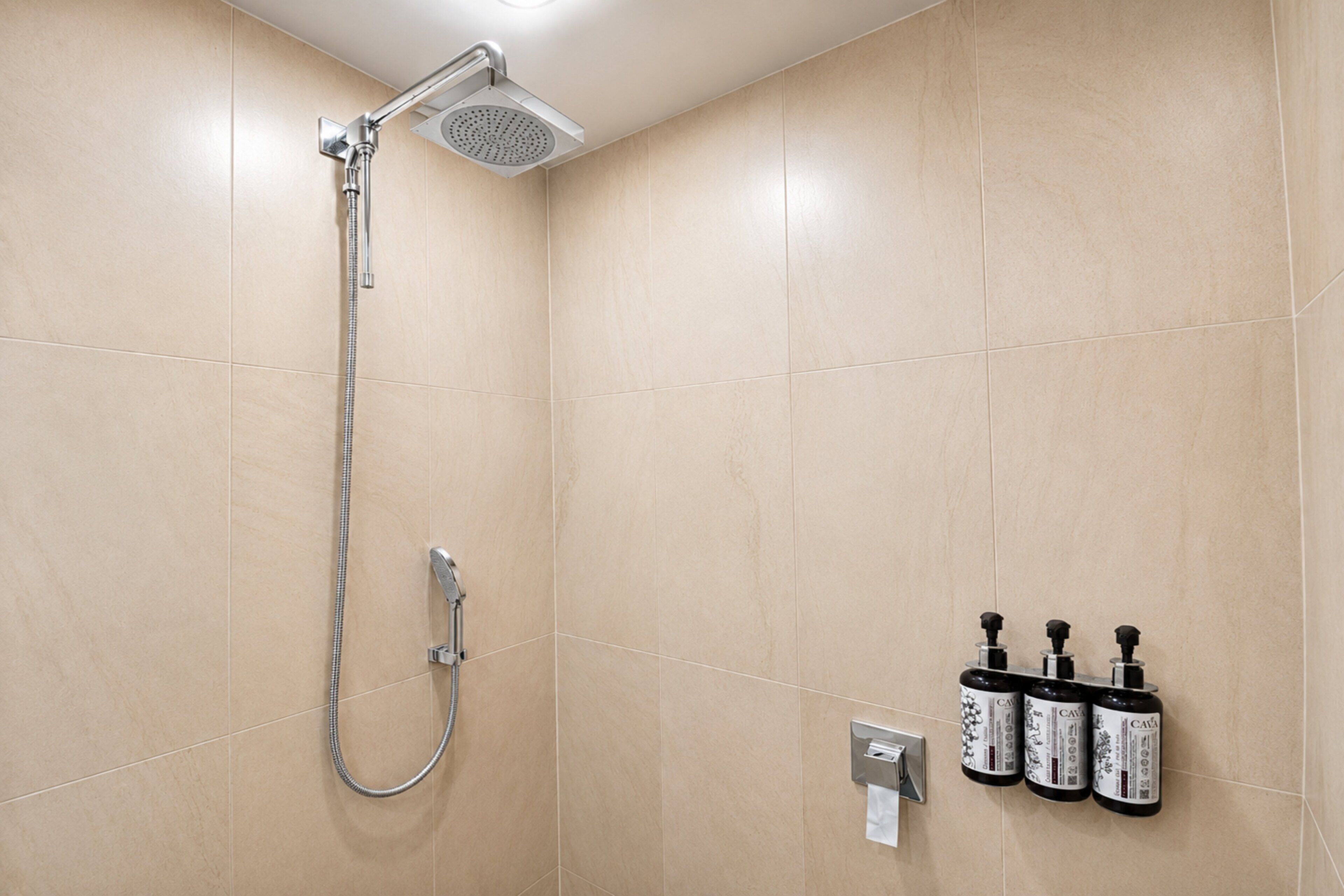 Une douche équipée d’un pommeau de douche à main et d’un robinet mural. Trois bouteilles de vin CAVA sont posées sur une étagère fixée au mur.