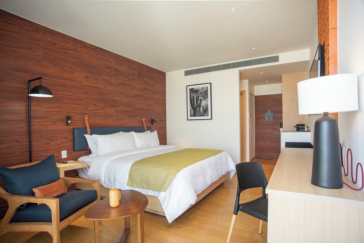 A modern hotel room with a wooden accent wall, a bed with white bedding, a desk with a lamp, and a chair.