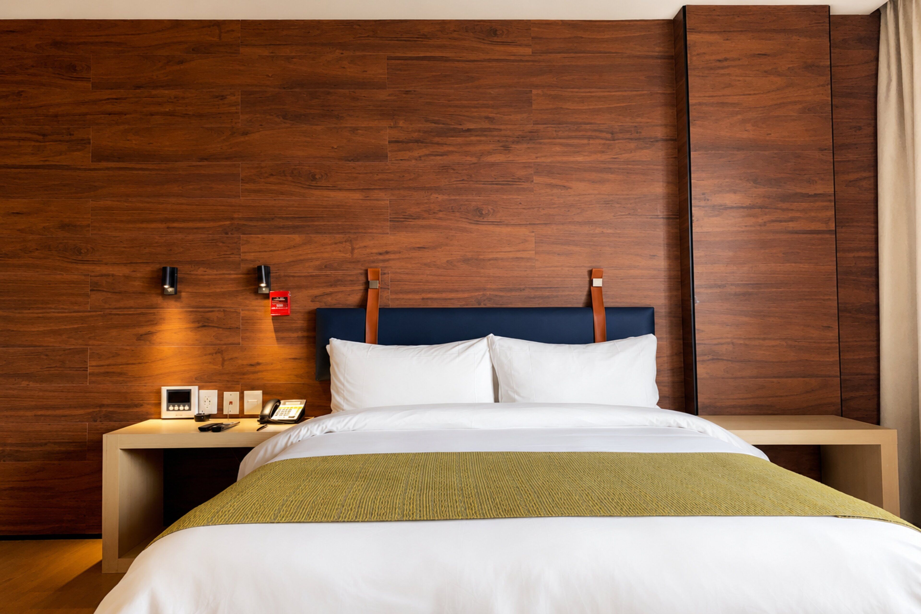 A hotel room with a wooden headboard, a bed with white bedding, a bedside table with a phone and a lamp, and a wall-mounted light fixture.