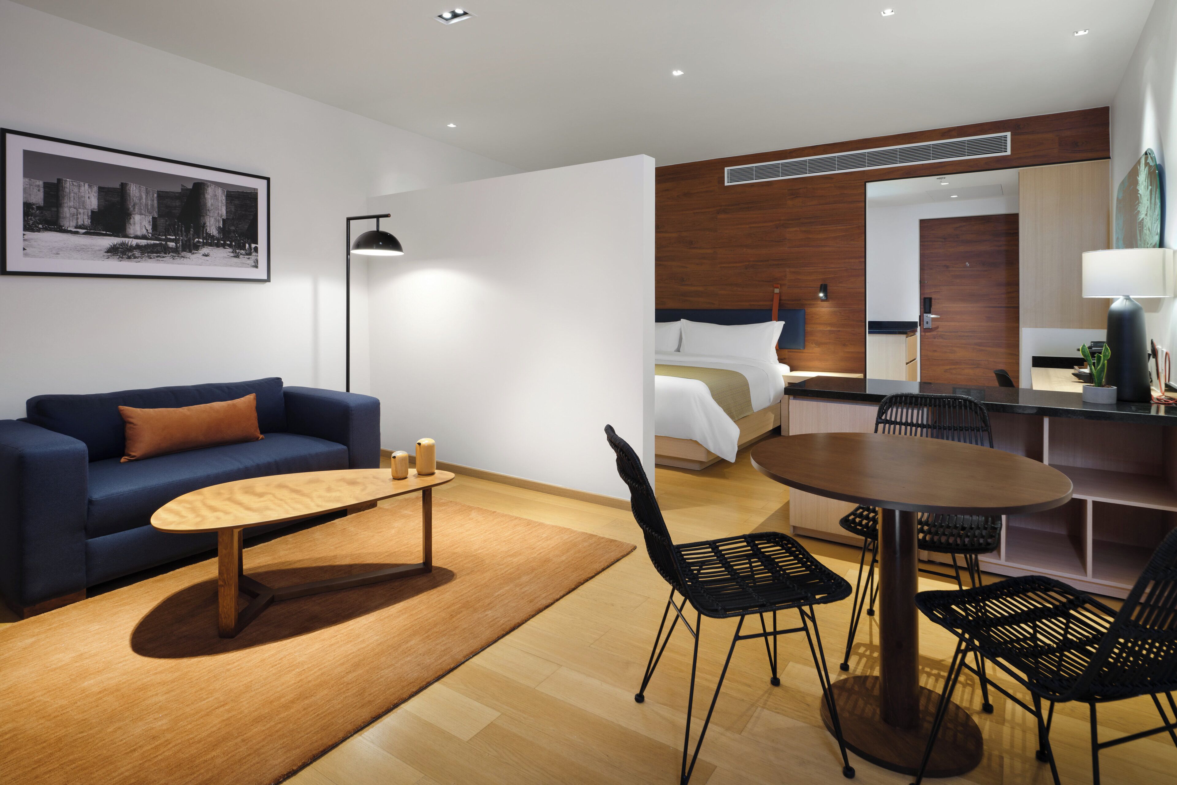 A modern hotel room with a sofa, coffee table, dining table, and chairs. There is a bed with a wooden headboard and a bedside table. A painting is on the wall, and a lamp is on the floor.