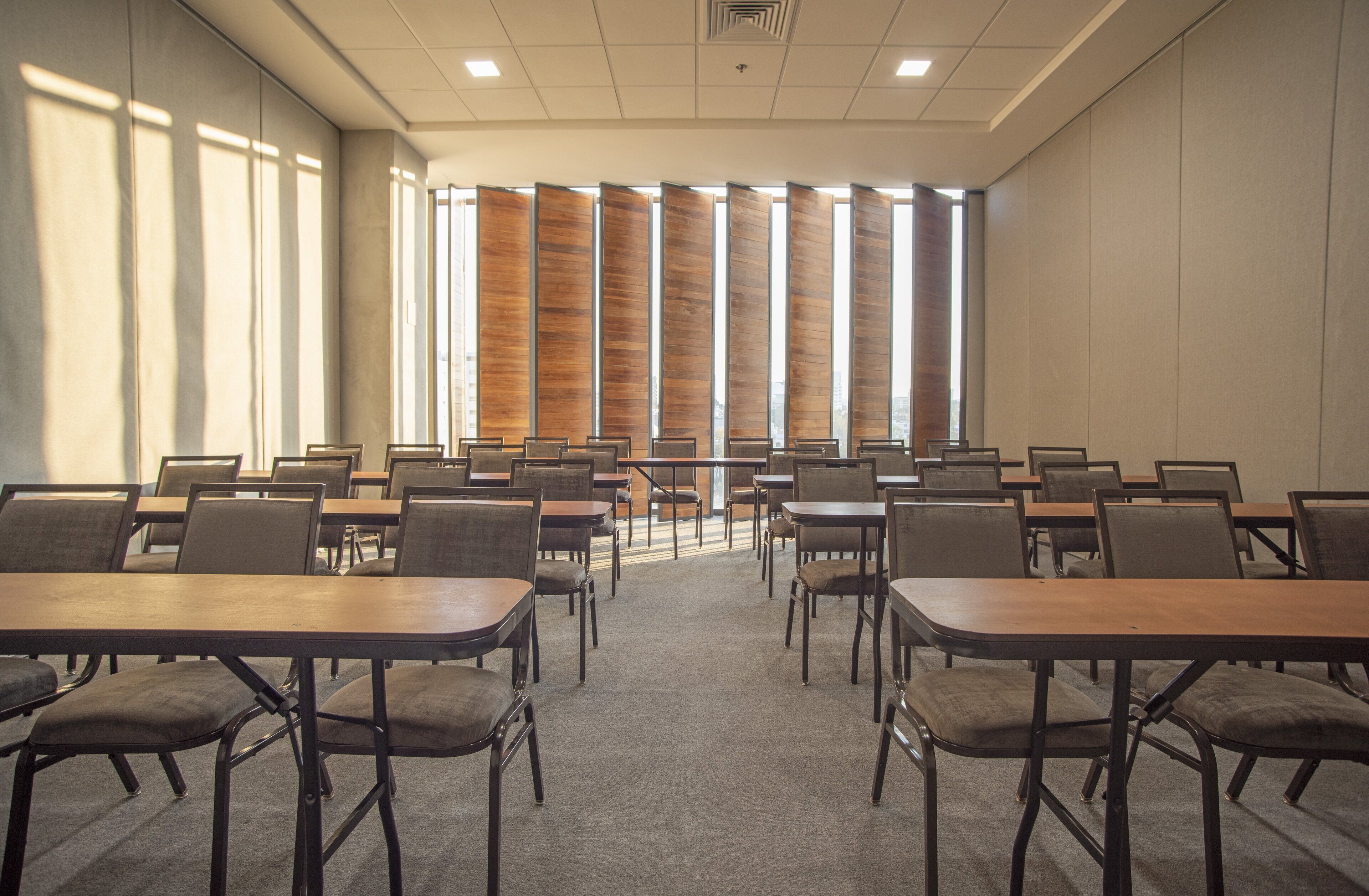 Un salón de conferencias con varias mesas y sillas, amplias ventanas con persianas y una decoración en tonos neutros.