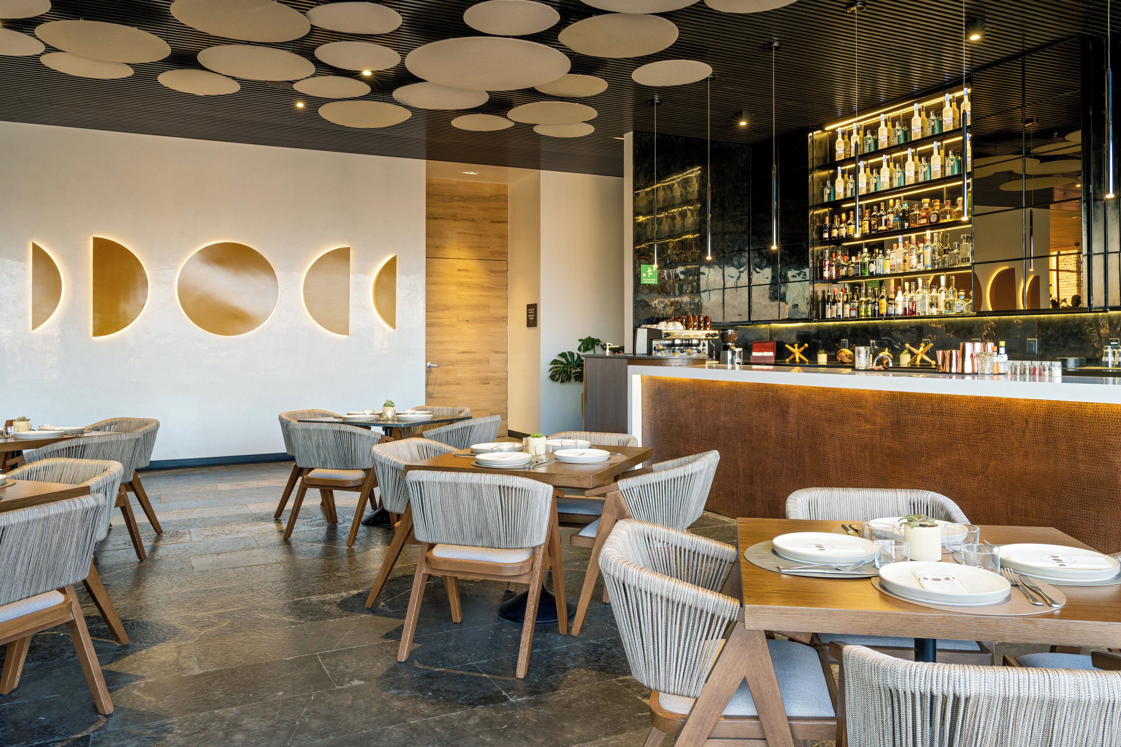 A modern restaurant interior with round tables, wicker chairs, a bar area with shelves of bottles, and a wooden accent wall.