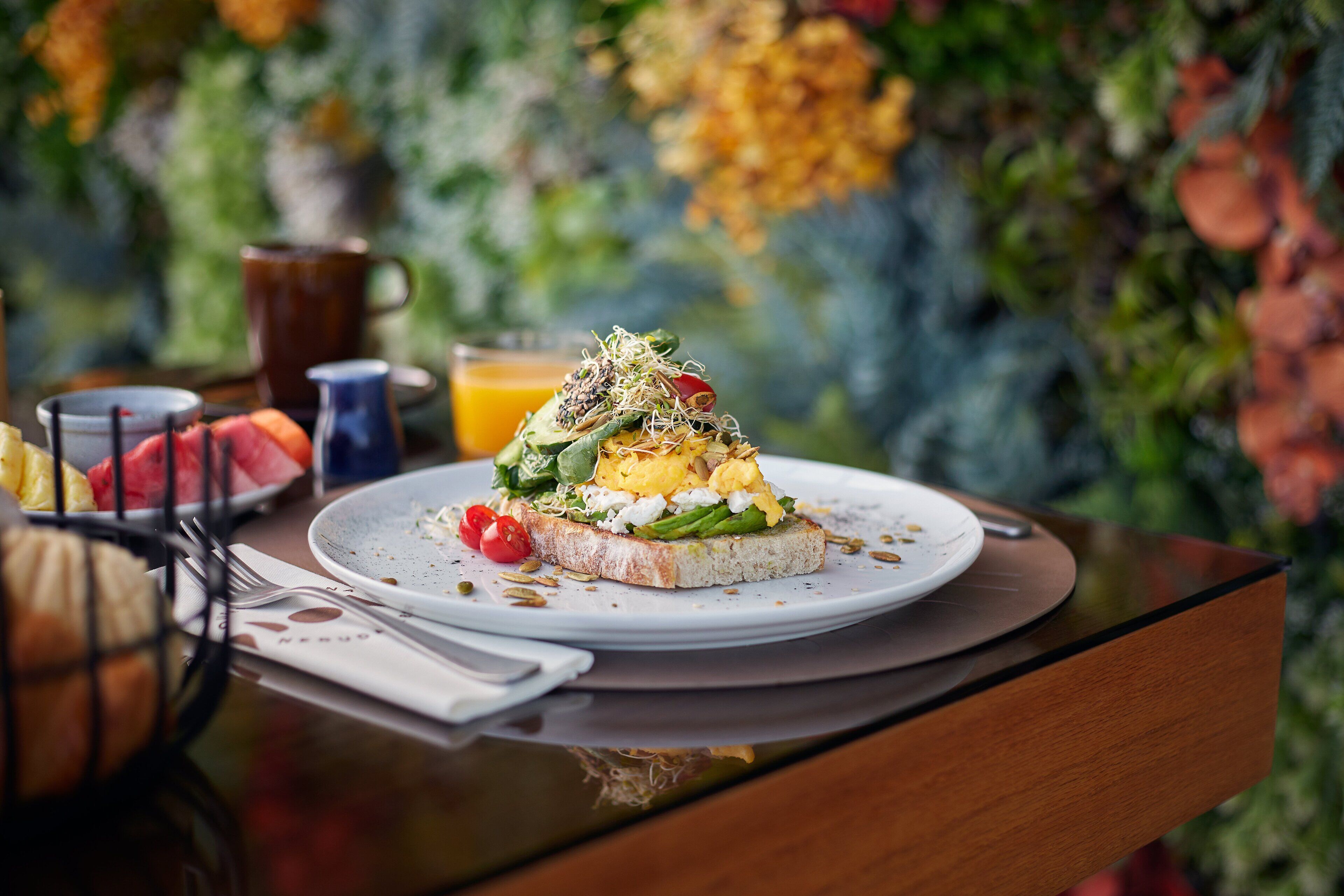 Een ontbijtsetting met een bord met toast belegd met diverse ingrediënten, een mok en een glas jus d'orange op een tafel met een houten blad.