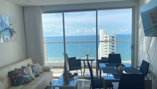 A modern living room with a large window overlooking the sea, a white sofa, a coffee table, and a dining area with chairs. - Beautiful condo with views of the beach (Cartagena)