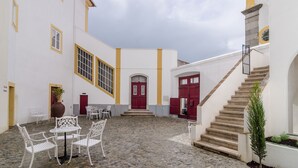 A courtyard with white walls, yellow trim, and red doors. There are white chairs and a table in the center. A staircase leads up to the entrance. - Casa Morgado Esporão (Évora)