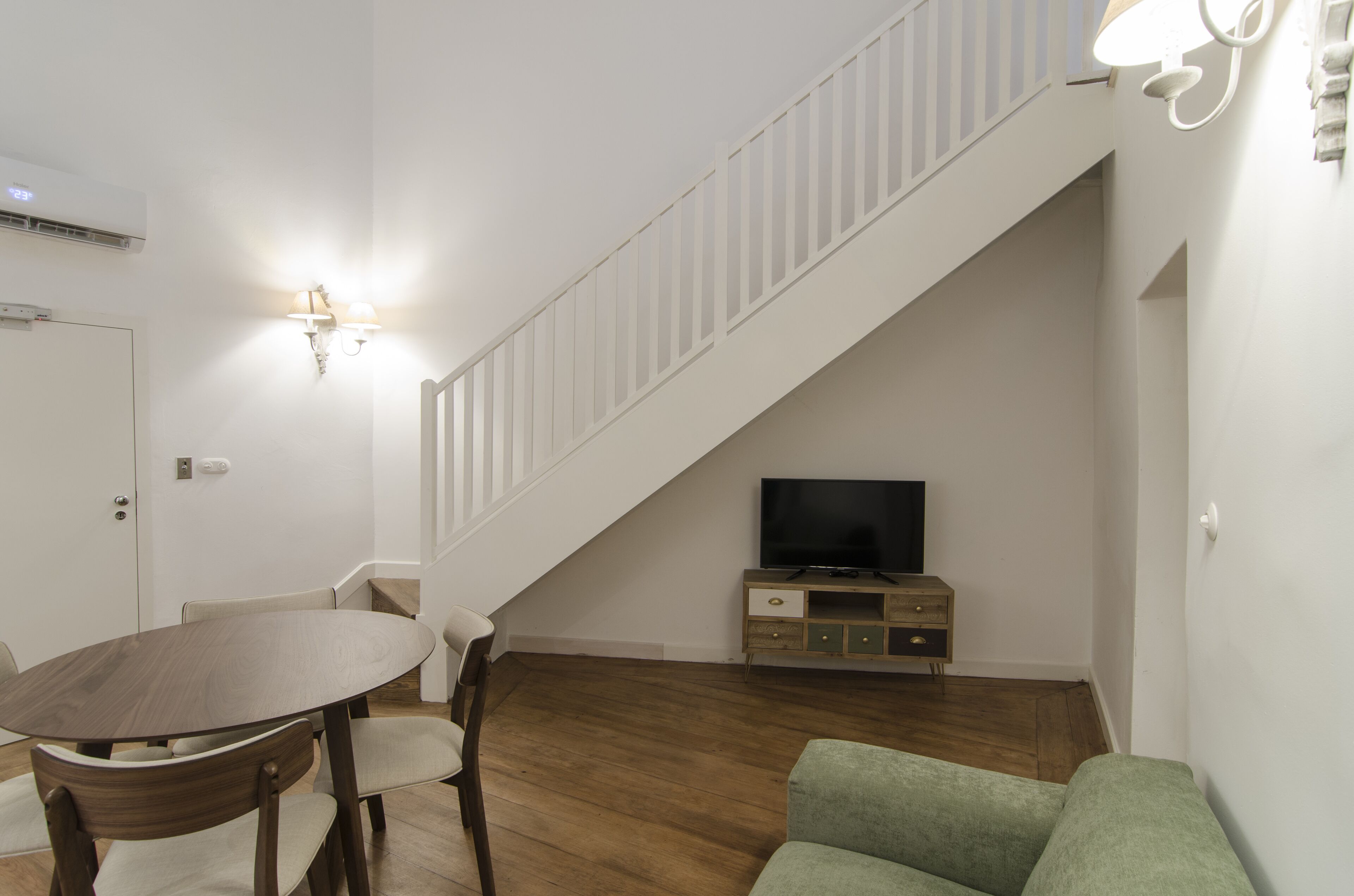 Una sala de estar con una mesa de comedor de madera, un sofá verde, un televisor sobre un mueble de madera y una escalera blanca.