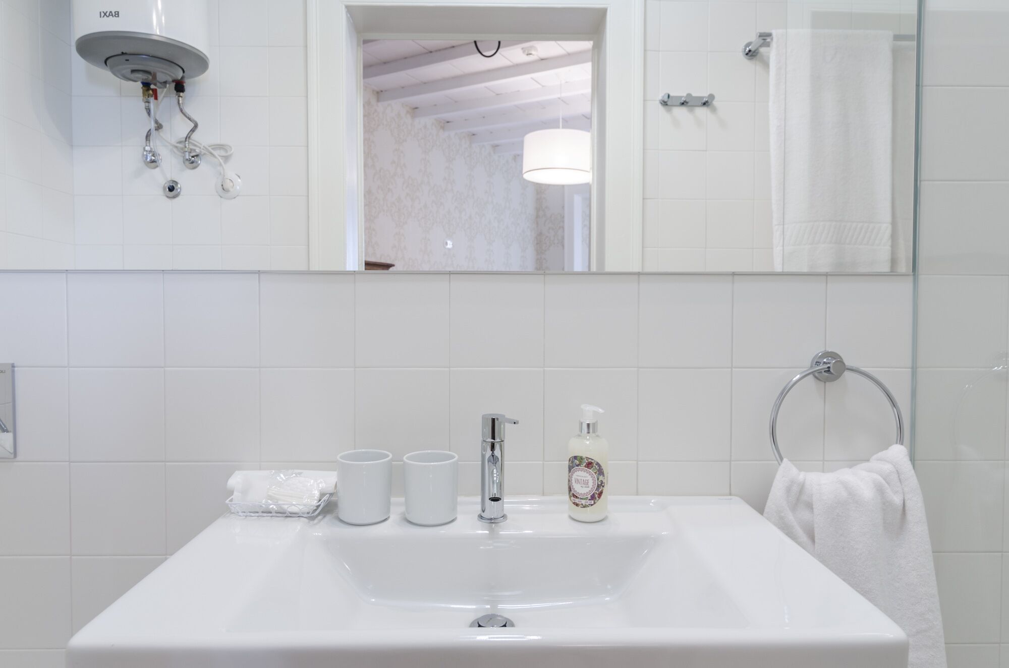 A bathroom with a white sink, two white cups, a soap dispenser, and a towel ring.