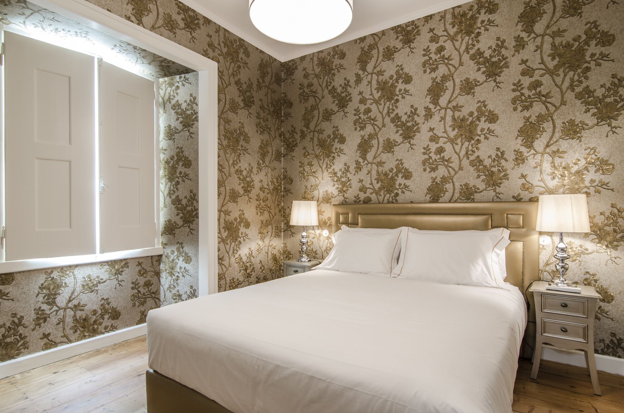 A bedroom with a large bed, two bedside tables with lamps, floral wallpaper, and a door with a white frame.