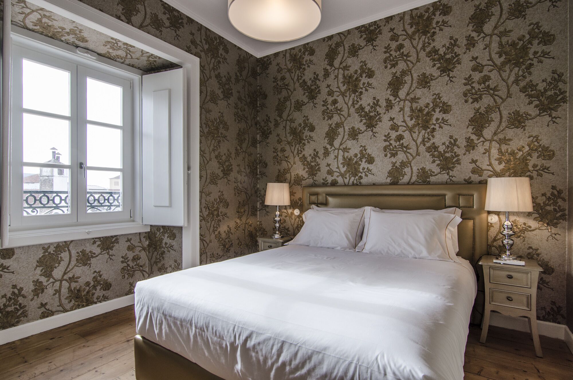 A bedroom with a large bed, two bedside tables with lamps, floral wallpaper, and a window with a view.