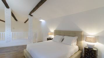 A bedroom with a large bed, white brick wall, wooden beams, and a nightstand with a lamp.