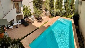 A rectangular swimming pool with a wooden deck and lounge chairs, surrounded by buildings and tall cypress trees. - Casa Morgado Esporão (Évora)