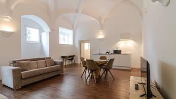 A spacious living area with a sofa, dining table, and chairs, a kitchen in the background, and a TV on a stand.