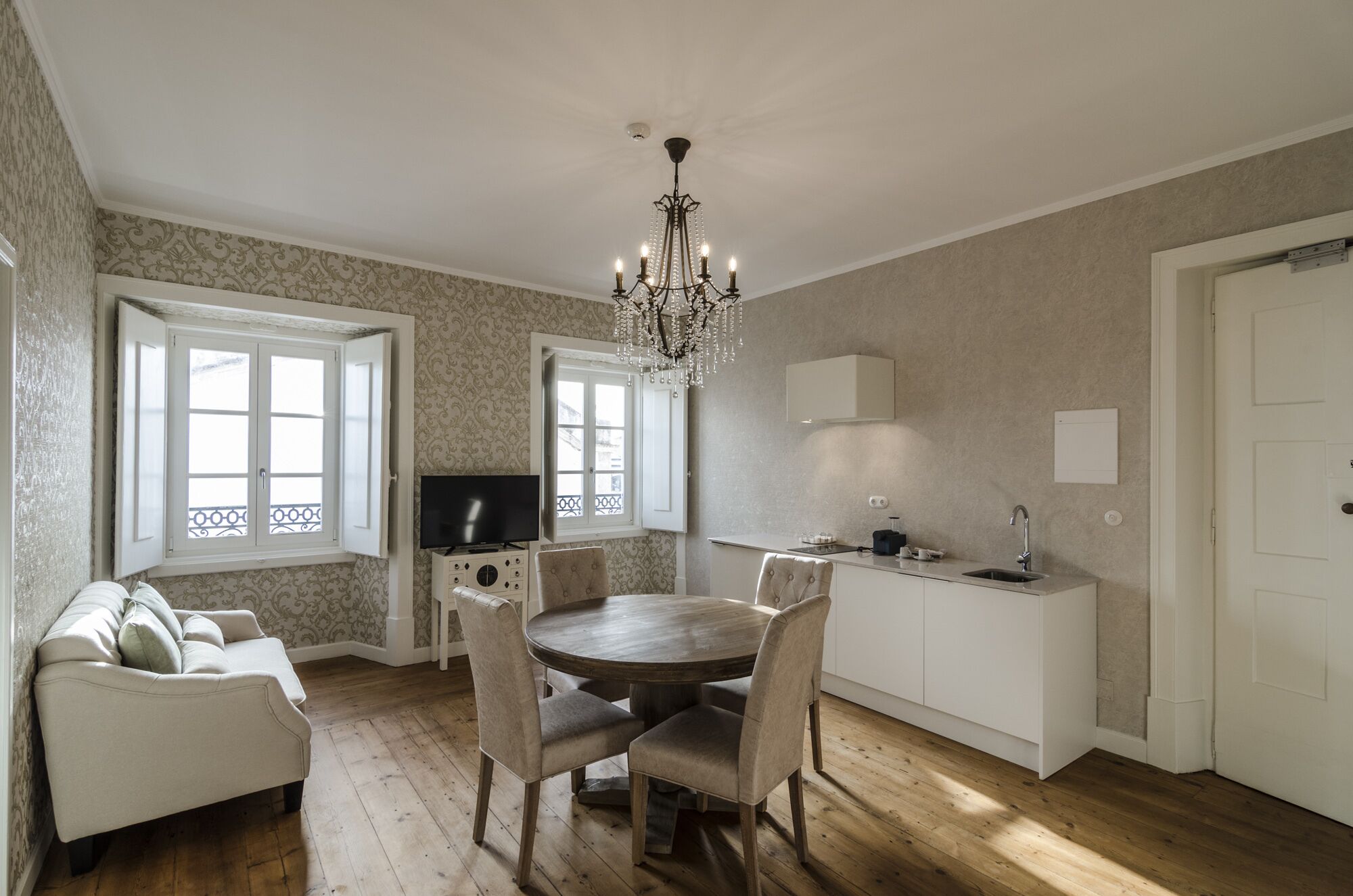 A dining area with a round table and chairs, a chandelier, a television, and a kitchen area with a sink and cabinets.