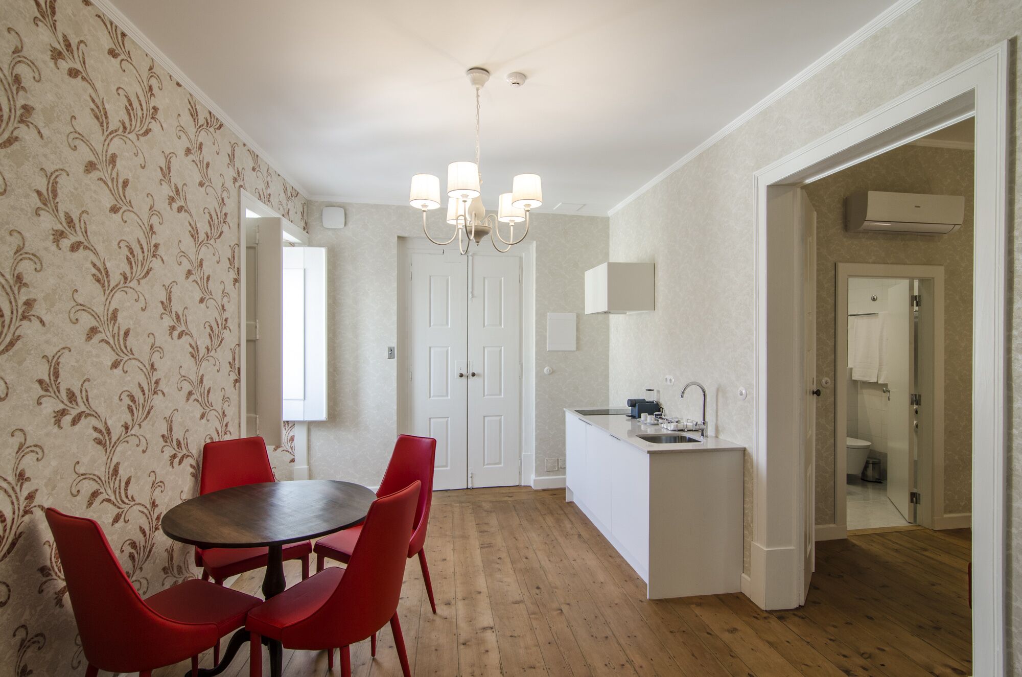 A compact kitchen with a central island, a dining area with red chairs, and a hallway leading to a bathroom.