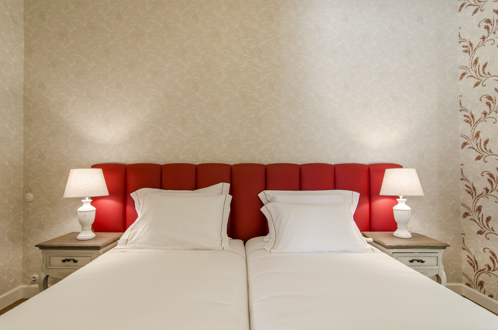 A double bed with white linens and red headboard, flanked by two bedside tables with lamps, in a room with patterned wallpaper.